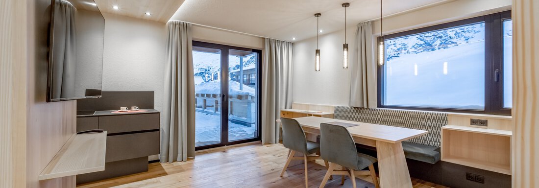 A modern living room with wooden floors and large windows. There is a dining area with a table and chairs as well as a television on the wall. - Hotel-Garni Pirchhütt - Obergurgl 
