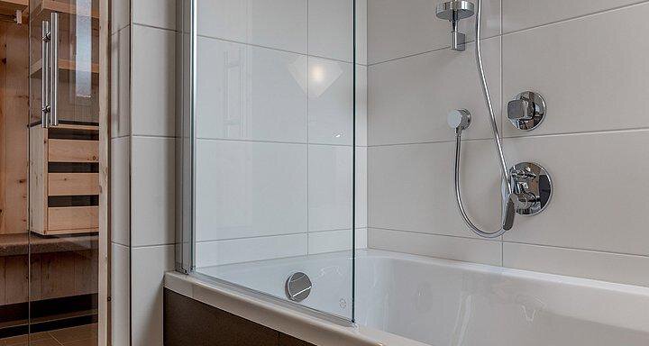 A modern bathroom with a bathtub and a shower. The walls are clad with white tiles and there are wooden details in the background. - Hotel-Garni Pirchhütt - Obergurgl 
