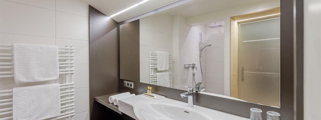 A modern bathroom with a large mirror and elegant fixtures. Towels and a towel rack are clearly visible, while the shower is positioned in the background. - Hotel-Garni Pirchhütt - Obergurgl 