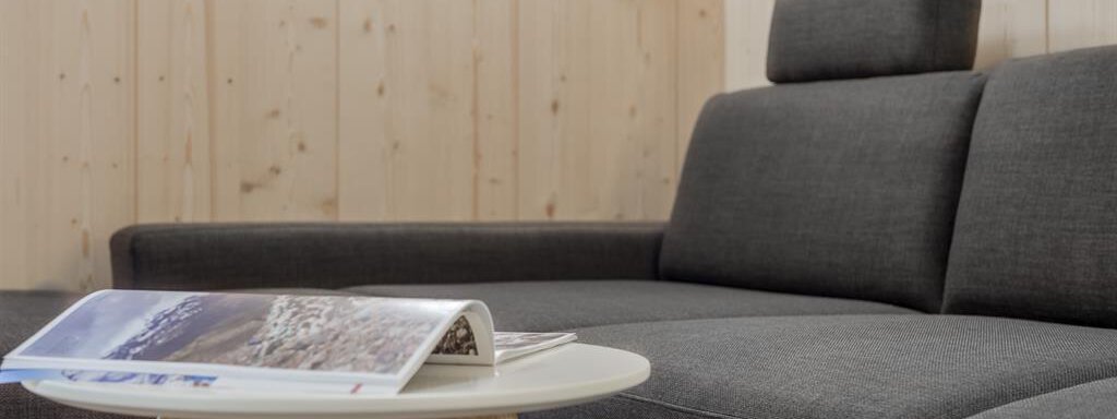 A modern living room with a gray couch and a small table. On the table lies a stack of magazines. - Hotel-Garni Pirchhütt - Obergurgl 