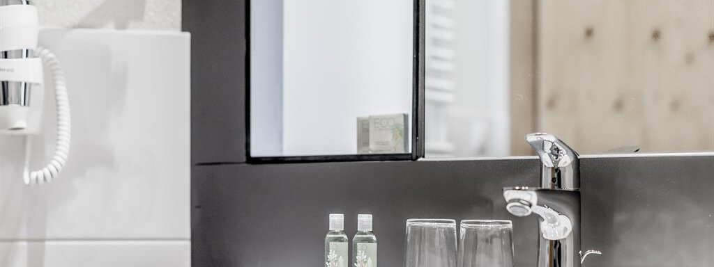 A modern bathroom with an elegant sink and a large mirror. Small bottles are placed next to two glasses. - Hotel-Garni Pirchhütt - Obergurgl 