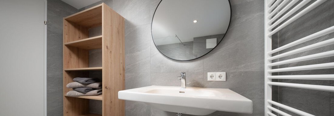 A modern bathroom with a round mirror and a wall-mounted sink. Next to the sink is a wooden shelf with towels. - Mountain Lodge Obergurgl - Obergurgl 