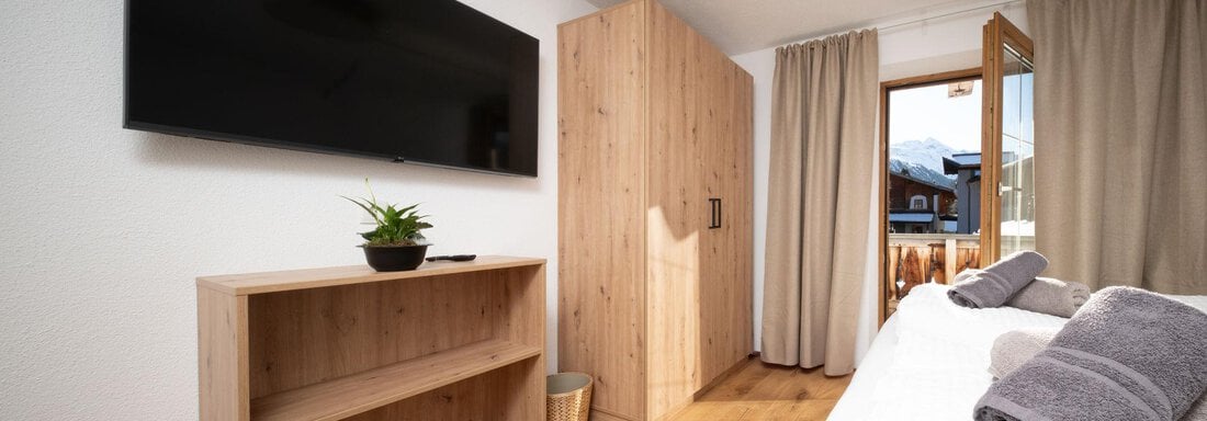 A modern bedroom with wooden furniture and a large TV on the wall. The windows let in plenty of natural light and offer a view outside. - Mountain Lodge Obergurgl - Obergurgl 