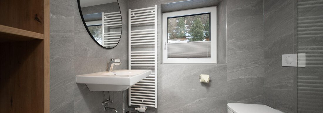 A modern bathroom with gray tiles and a large mirror. The amenities include a wall-mounted toilet and a sink with space for toiletries. - Mountain Lodge Obergurgl - Obergurgl 
