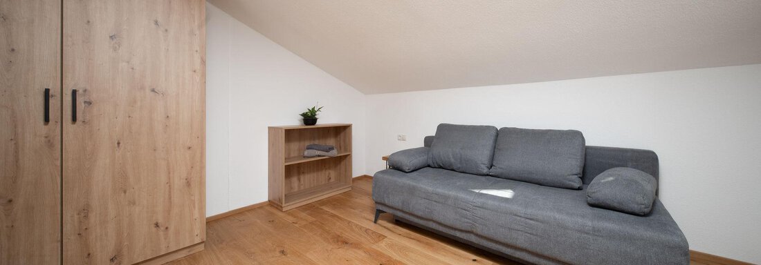 A cozy attic room with a gray sofa and a wooden wardrobe. The floor is made of light wood and there is a small plant on the shelf. - Mountain Lodge Obergurgl - Obergurgl 