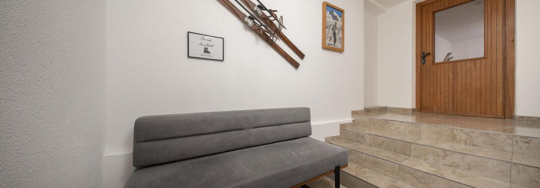 A modern entrance area with a gray sofa and a staircase. Old skis and a framed picture are hanging on the wall. - Mountain Lodge Obergurgl - Obergurgl 