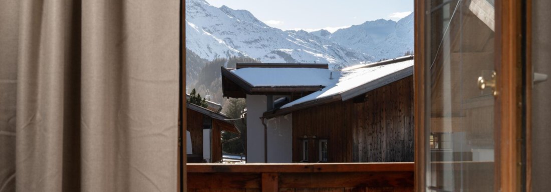 View from a window overlooking snow-covered mountains and wooden chalets. The image conveys a cozy and alpine atmosphere. - Mountain Lodge Obergurgl - Obergurgl 