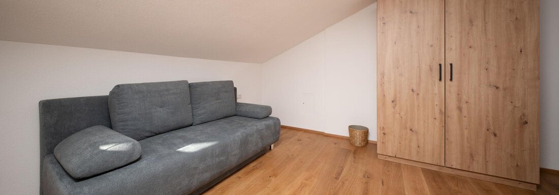 A cozy room with a gray sofa and a wooden wardrobe. The floor is made of light wood and the walls are plain and white. - Mountain Lodge Obergurgl - Obergurgl 