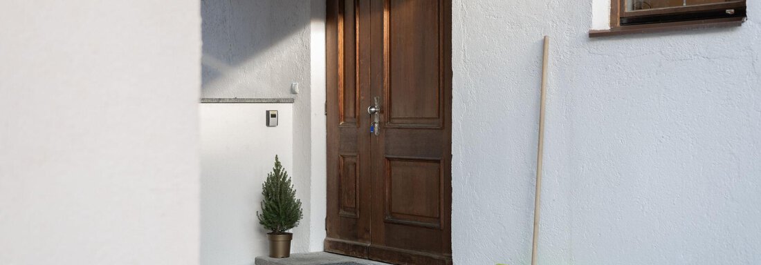 A wooden door with a green plant pot stands in front of a light wall area. Next to the door, a green shovel lies on the ground. - Mountain Lodge Obergurgl - Obergurgl 