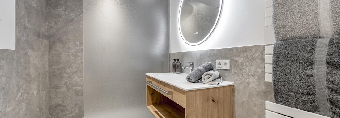 A modern bathroom with a glass shower and a wooden sink. Towels are neatly folded and a round mirror light provides pleasant illumination. - Peak Gurgl - Obergurgl 