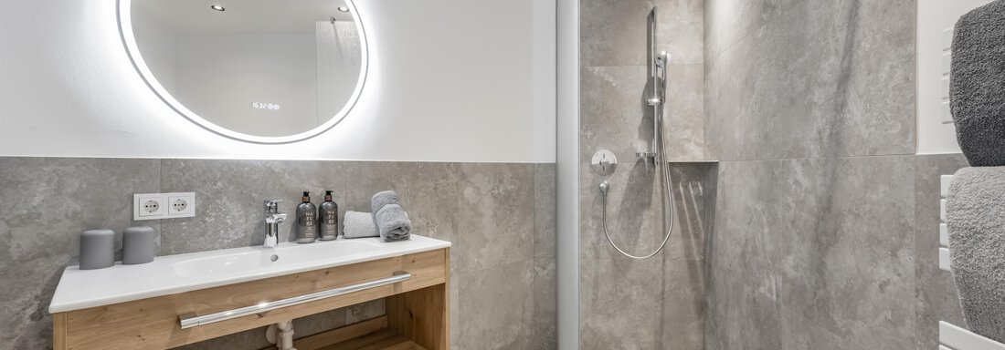 A modern bathroom with an elegant shower and a wooden sink cabinet. The round mirror with lighting creates a stylish atmosphere. - Peak Gurgl - Obergurgl 