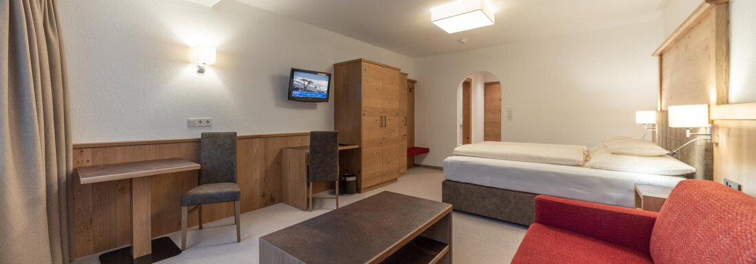Ein modernes Hotelzimmer mit einem Doppelbett, einem roten Sofa und einem Schreibtisch. Die Wände sind hell und der Raum ist gut beleuchtet. - Pension Hohenfels - Obergurgl 