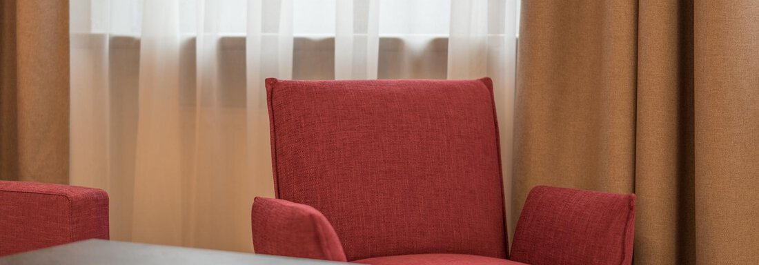 Ein gemütlicher roter Sessel steht neben einem Holztablett. Im Hintergrund sind helle Vorhänge zu sehen. - Pension Hohenfels - Obergurgl 