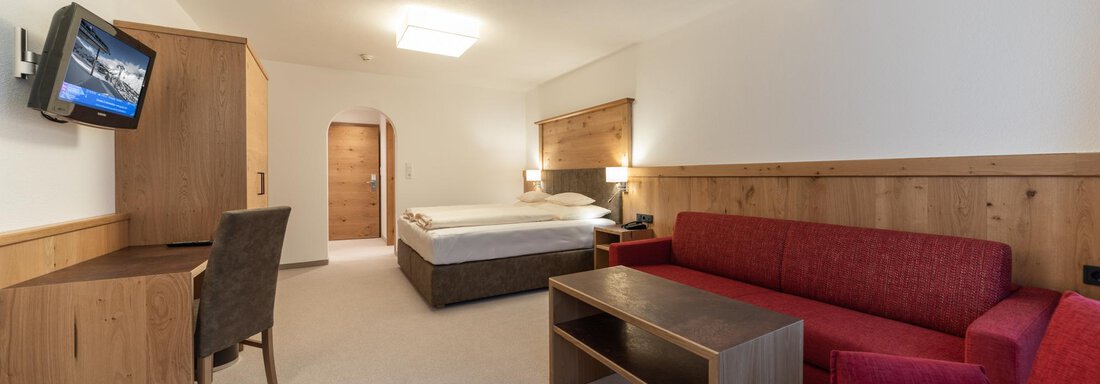 Ein modernes Hotelzimmer mit einem Doppelbett, einem roten Sofa und einem Schreibtisch. Die Wände sind in neutralen Farben gehalten, und es gibt einen TV an der Wand. - Pension Hohenfels - Obergurgl 