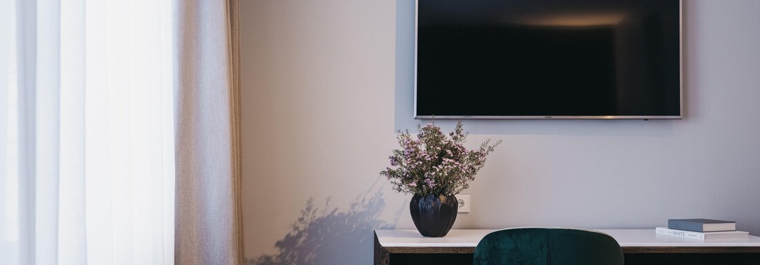 A modern room with a desk and a television on the wall. A vase with flowers sits on the table and a window with curtains lets light in. - The Crystal VAYA Unique - Obergurgl 