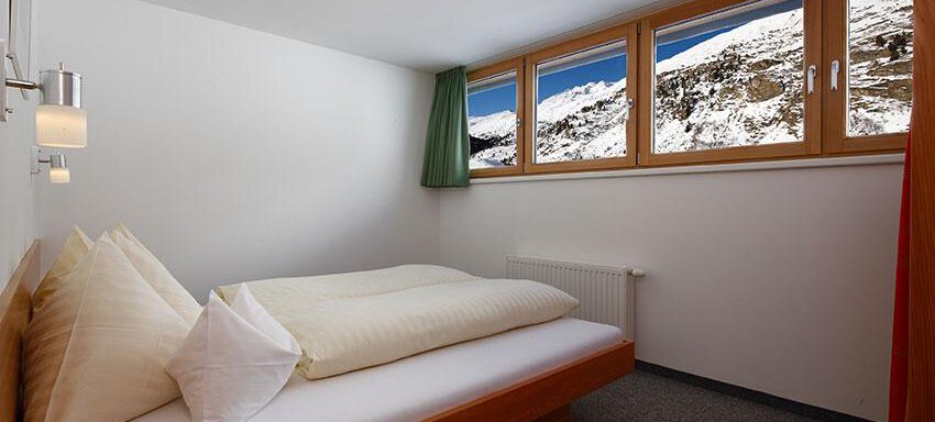 A cozy bedroom with a double bed and two windows that offer a view of snow-covered mountains. The walls are painted light, and the furnishings are simple and modern. - Ferienwohnungen Broser - Obergurgl 