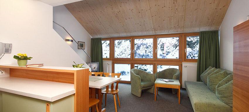 Wohnküche A cozy living room with a wooden ceiling and large windows. There is a seating area with a sofa and a dining area. - Ferienwohnungen Broser - Obergurgl