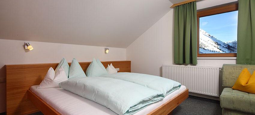 A modern bedroom with a large bed and bright, cozy pillows. Through the window, you can see the mountains and the clear sky. - Ferienwohnungen Broser - Obergurgl 
