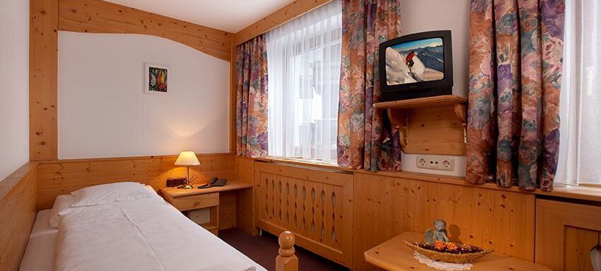 A cozy single room with wooden paneling and colorful curtains. There is a bed, a table, and a television. - Haus Martinshof - Obergurgl 