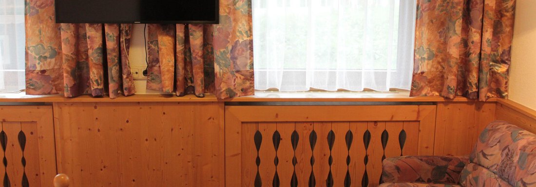 A cozy living room with a television on the wall and a colorful sofa. The windows are adorned with curtains and let in bright light. - Haus Martinshof - Obergurgl 
