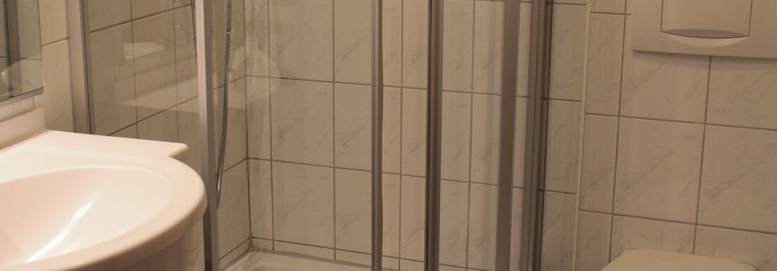 A simple bathroom with a shower cabin, a sink, and a toilet. The walls are tiled with light-colored tiles. - Haus Martinshof - Obergurgl 