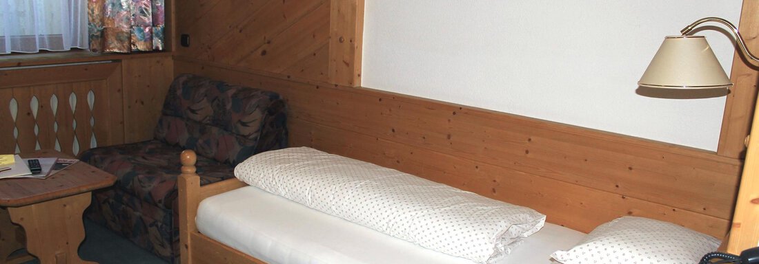 A simple room with wooden walls and a single bed. There is an armchair, a table, and a window with curtains. - Haus Martinshof - Obergurgl 
