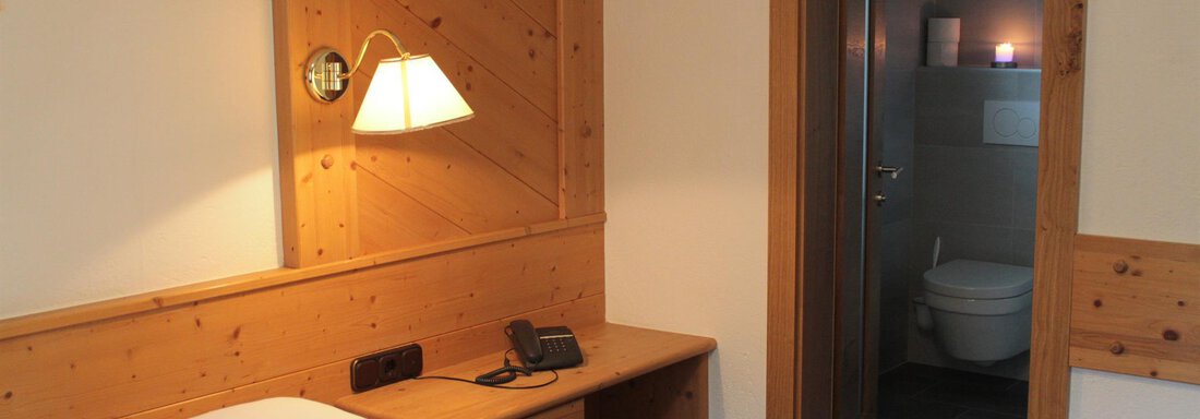 A simple room with wooden paneling and a single bed. In the background, a bathroom with a toilet is visible. - Haus Martinshof - Obergurgl 