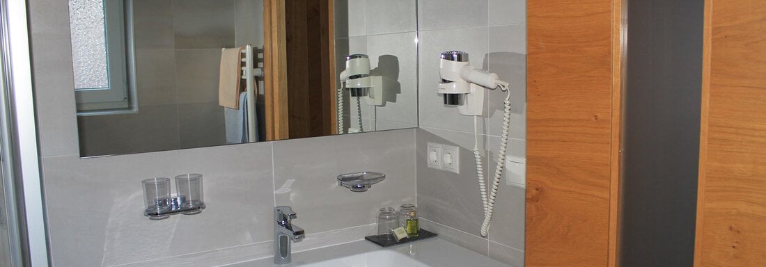 A modern bathroom with a vanity, a mirror, and towels. The walls are light tiled and the furniture has wood details. - Haus Martinshof - Obergurgl 