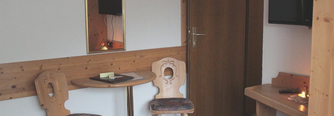 A simple room with wooden furniture, two chairs, and a small table. A television hangs on the wall and a mirror provides light. - Haus Martinshof - Obergurgl 
