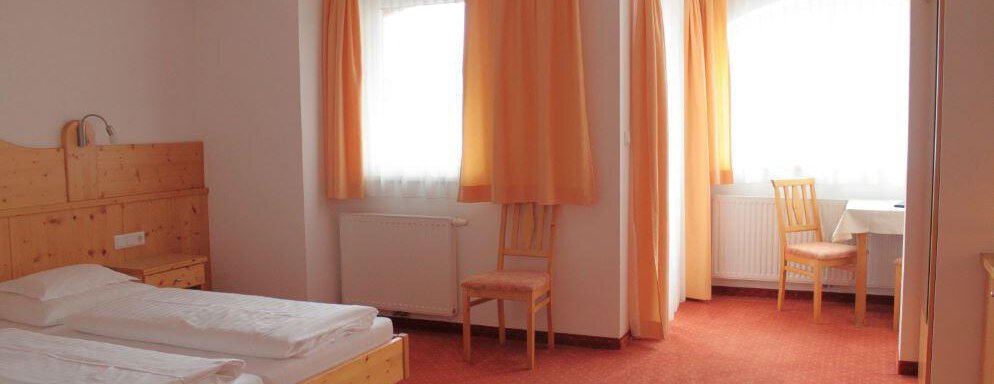 A simple bedroom with a double bed and wooden furniture. There are large windows with orange curtains and a workspace with a table and a chair. - Haus Martinshof - Obergurgl 