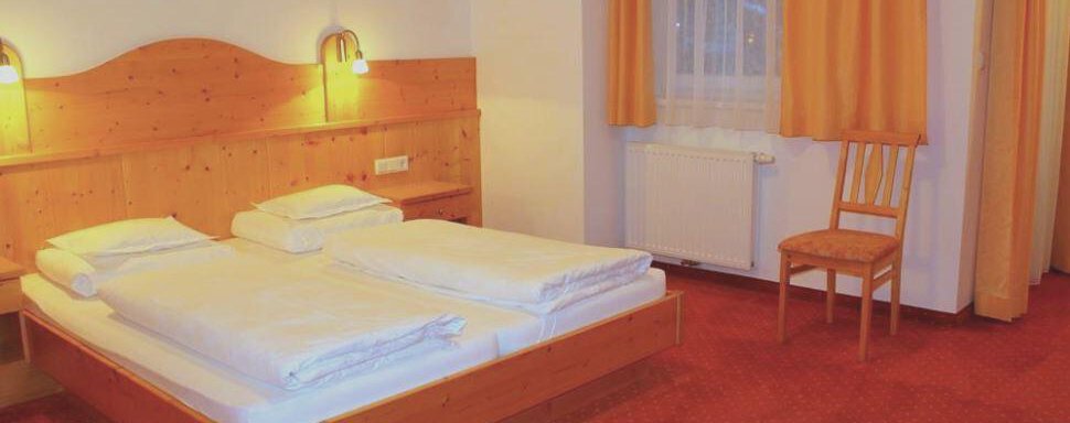 A cozy bedroom with a double bed and wooden furniture. The walls are painted light, and there are large windows with curtains in bright orange. - Haus Martinshof - Obergurgl 