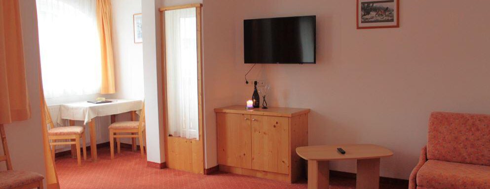 A spacious room with a TV, a table, and chairs. The walls are light, and there are large windows with curtains. - Haus Martinshof - Obergurgl 