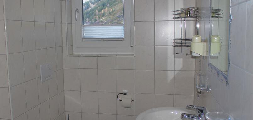 A simple bathroom with white tiles and natural light. It includes a toilet, a hand basin, and a mirror. - Haus Martinshof - Obergurgl 