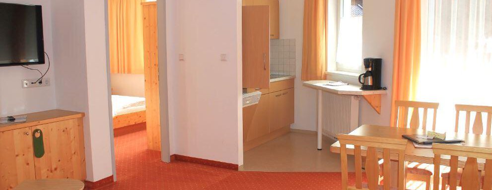 A bright living and dining room with a table, chairs, and a television. The room has large windows with curtains in warm colors. - Haus Martinshof - Obergurgl 