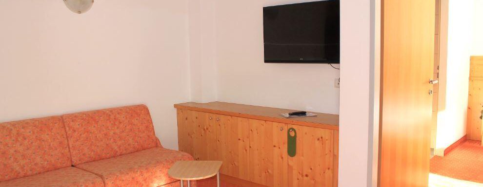 A cozy room with an orange sofa and a TV on the wall. The floor is covered with a red carpet and a door leads to another room. - Haus Martinshof - Obergurgl 
