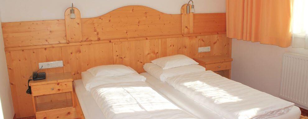 A simple room with two single beds and wooden furniture. The walls are light, and there are large windows with curtains. - Haus Martinshof - Obergurgl 
