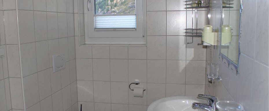 A simple bathroom with white tiles and a window. It contains a sink, a toilet, and shelves for toiletries. - Haus Martinshof - Obergurgl 