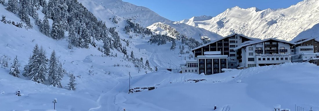 A snowy mountain landscape with a modern hotel. The sky is clear and blue, and the mountains rise majestically in the background. - Haus Verwall Appartements - Obergurgl 