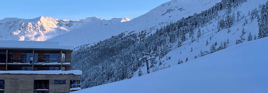 A snowy mountain landscape with tall mountains in the background. In the foreground, a modern building is visible. - Haus Verwall Appartements - Obergurgl 