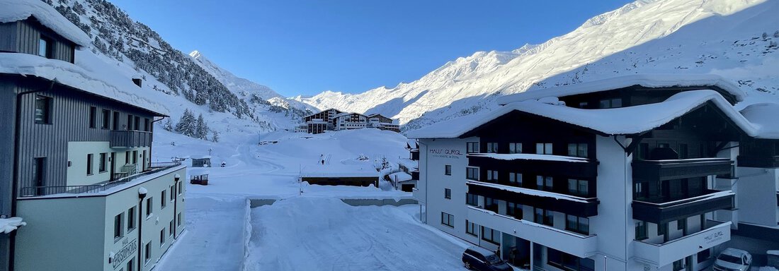 A snowy landscape with modern buildings in the foreground. In the background, snow-covered mountains are visible under a clear blue sky. - Haus Verwall Appartements - Obergurgl 