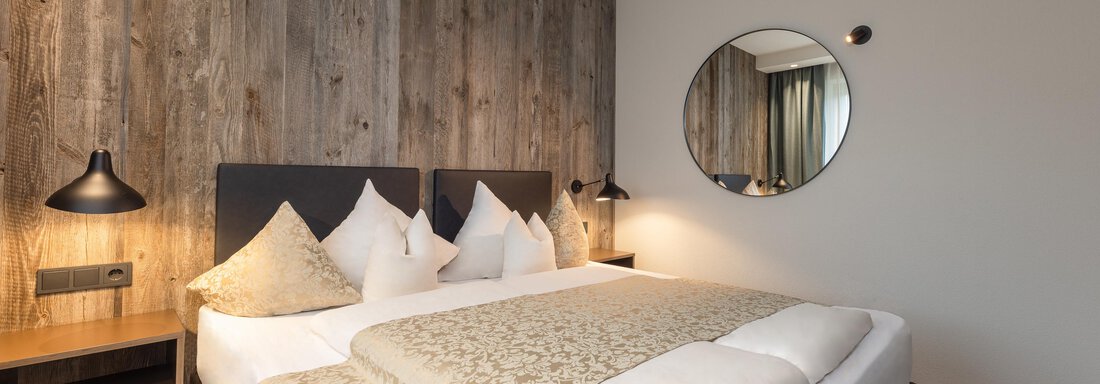 A modern bedroom with a cozy double bed and elegant wood paneling. The round wall mirror and warm lighting create an inviting atmosphere. - Haus Verwall Appartements - Obergurgl 