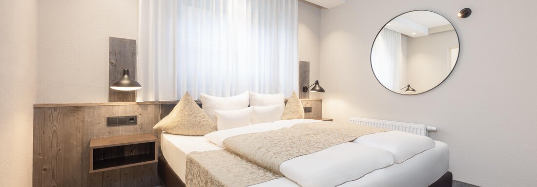 The image shows a modern bedroom with a large bed and bright decor. There is a round mirror and gentle lighting that creates a cozy atmosphere. - Haus Verwall Appartements - Obergurgl 