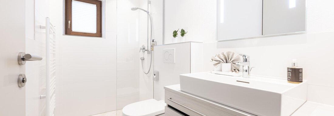A modern bathroom with white wall and floor colors. There is a shower, a sink, and a large mirror. - Haus Verwall Appartements - Obergurgl 