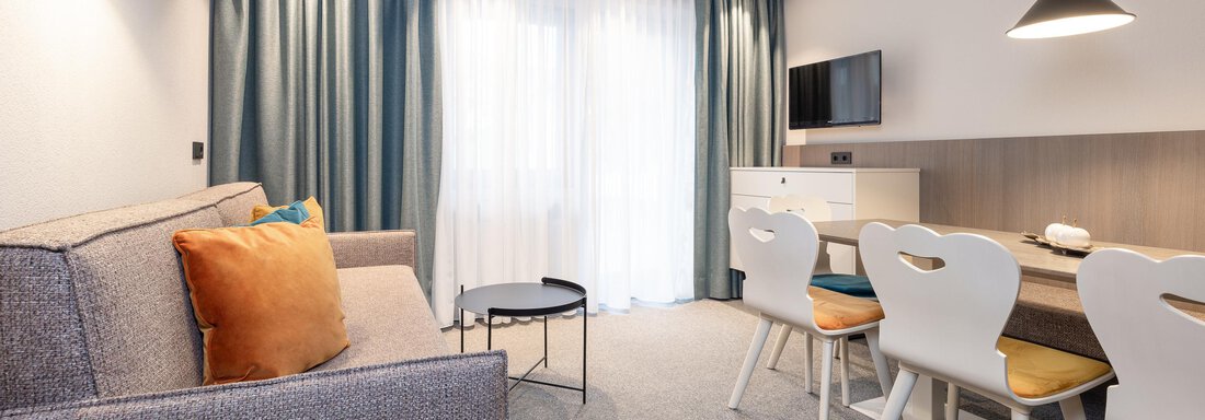 A modern living room with a gray sofa and a small table. There is a dining area with light chairs and a TV on the wall. - Haus Verwall Appartements - Obergurgl 