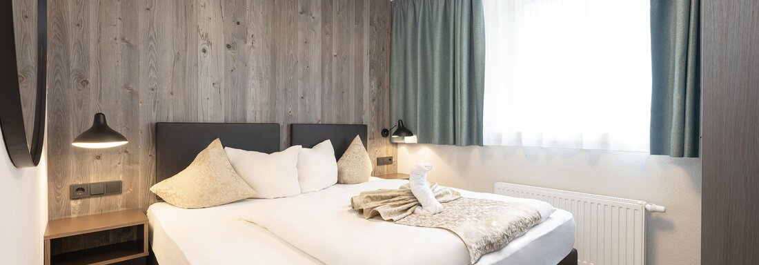 A modern bedroom with a large bed and wood paneling. The floor-to-ceiling windows allow plenty of daylight in. - Haus Verwall Appartements - Obergurgl 