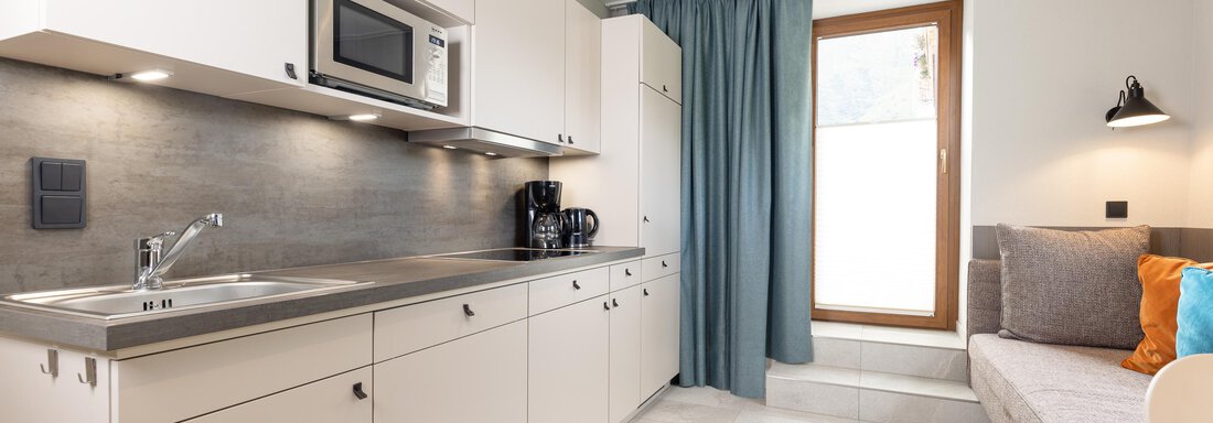 A modern kitchen with white cabinets and stainless steel appliances.  
Next to the kitchen, there is a cozy seating area with a window and blue curtains. - Haus Verwall Appartements - Obergurgl 