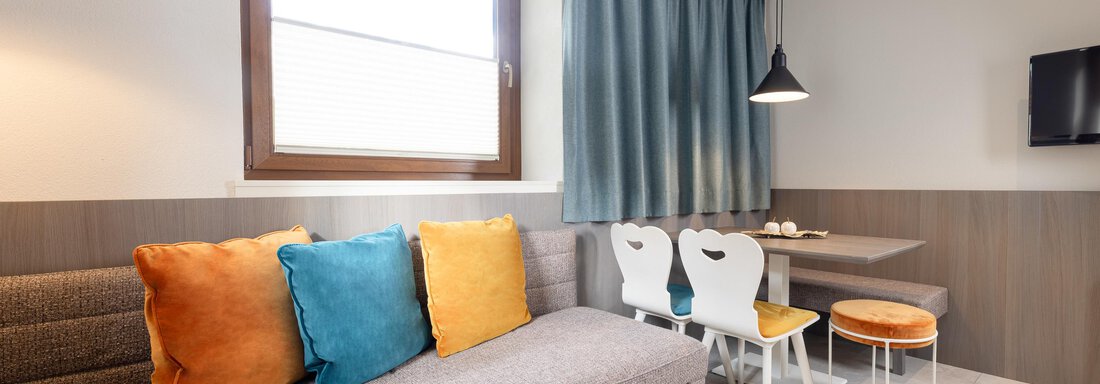 A modern living room with a gray couch and colorful cushions. A dining table with chairs and a large window creates a bright, inviting atmosphere. - Haus Verwall Appartements - Obergurgl 