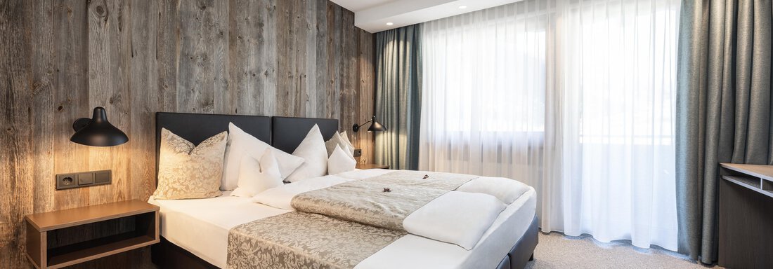 A modern bedroom with a large bed and a wooden wall.  
A lot of light pours through the windows, and the room feels inviting. - Haus Verwall Appartements - Obergurgl 