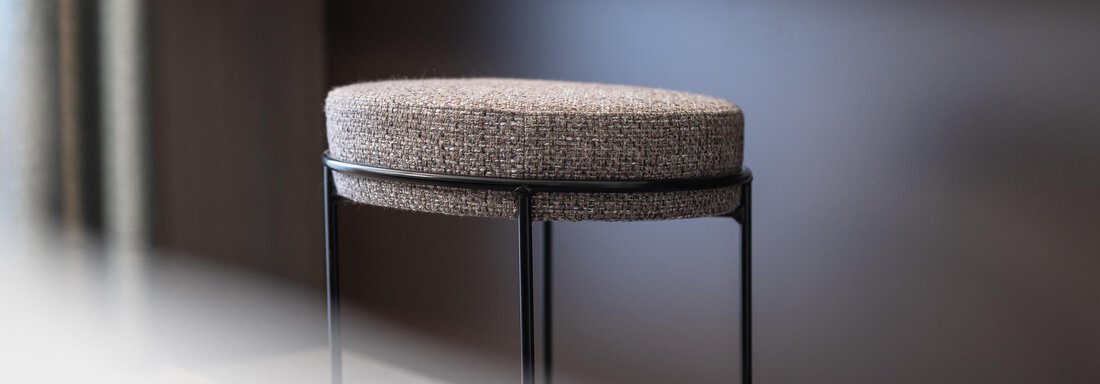 An elegant stool with a soft, patterned cover is placed in front of a plain wall. The simple atmosphere is complemented by a modern shelf. - Haus Verwall Appartements - Obergurgl 