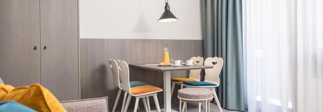 A modern living and dining room with a table, chairs, and a cozy couch. Bright curtains and decorative elements create an inviting atmosphere. - Haus Verwall Appartements - Obergurgl 
